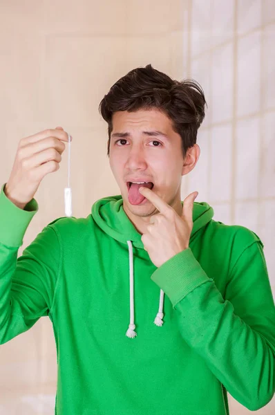 Stilig man med en äcklad ansikte håller en menstruation bomull tampong, gör ett tecken med fingret inuti munnen, iklädd en grön hoodie i en suddig bakgrund — Stockfoto