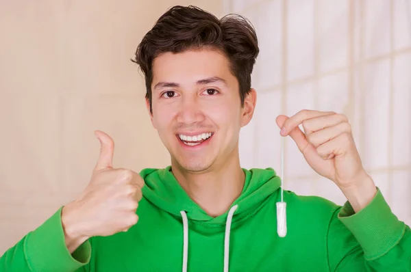 Guapo hombre sonriente con una cara feliz sosteniendo un tampón de algodón menstruación con el pulgar hacia arriba, aprobando el uso de tampones, usando una sudadera con capucha verde en un fondo borroso —  Fotos de Stock