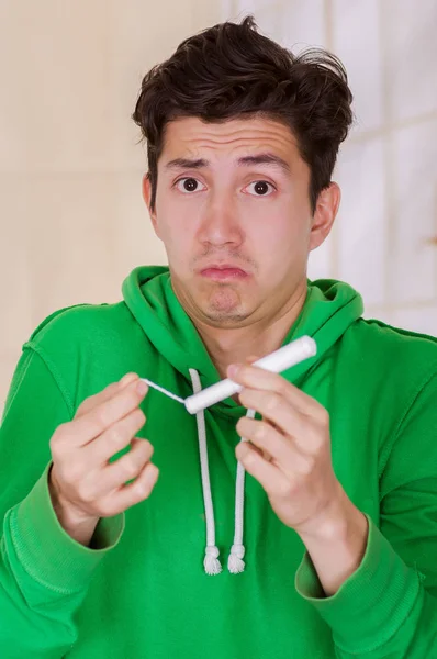 Hombre guapo con la cara perdida sosteniendo un tampón de algodón de menstruación, con una sudadera verde en un fondo borroso — Foto de Stock