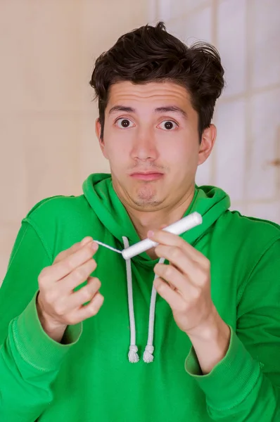 Hombre guapo con una cara divertida sosteniendo un tampón de algodón de menstruación, con una sudadera con capucha verde en un fondo borroso — Foto de Stock