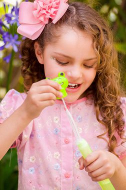 Bir yaz niteliğine sabun köpüğü ile oynarken, bir bulanık doğa arka planda kafasının içinde bir pembe elbise ve çiçek giyen mutlu küçük kıvırcık kız