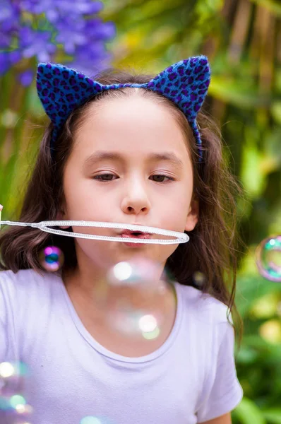 Happy little girl playing with soap bubbles on a summer nature, wearing a blue ears tiger accessories over her head in a blurred nature background