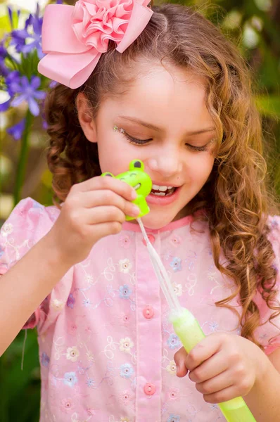 Happy little curly girl playing with soap bubbles on a summer nature, wearing a pink dress and flower in her head, in a blurred nature background — Stock Photo, Image
