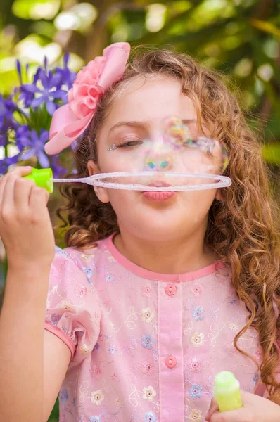 Happy little curly girl playing with soap bubbles on a summer nature, wearing a pink dress and flower in her head, in a blurred nature background — Stock Photo, Image