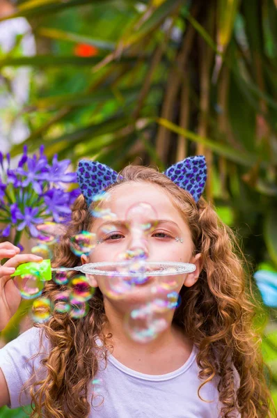 Happy little curly girl playing with soap bubbles on a summer nature, wearing a blue ears of tiger accessories over her head in a blurred nature background — Stock Photo, Image