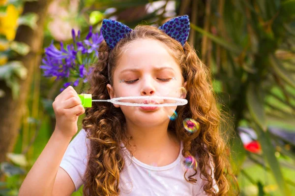 Glückliches kleines lockiges Mädchen, das mit Seifenblasen in der sommerlichen Natur spielt und ein blaues Ohr mit Tiger-Accessoires über dem Kopf in einem unscharfen Naturhintergrund trägt — Stockfoto