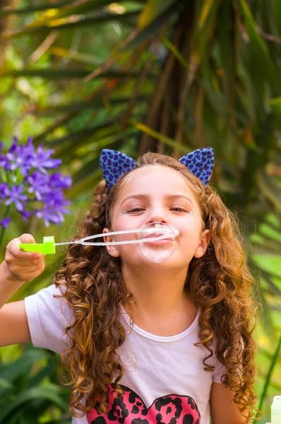 Glückliches kleines, lockig lächelndes Mädchen, das mit Seifenblasen in der sommerlichen Natur spielt und ein blaues Ohr mit Tiger-Accessoires über dem Kopf in einem verschwommenen Naturhintergrund trägt — Stockfoto