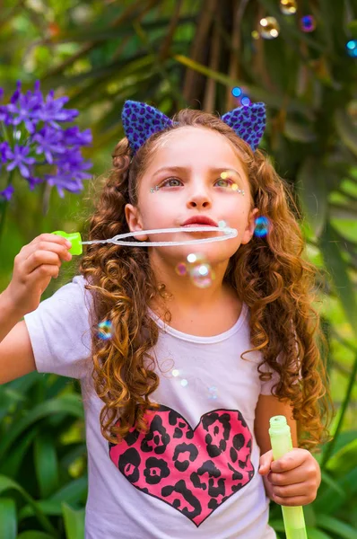Happy little curly girl playing with soap bubbles on a summer nature, wearing a blue ears of tiger accessories over her head in a blurred nature background — Stock Photo, Image