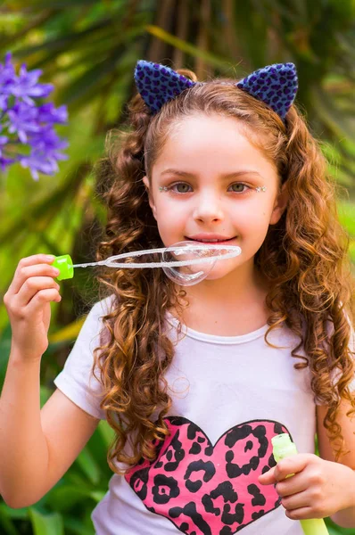 Blij krullend meisje spelen met zeepbellen op een zomer natuur, het dragen van een blauwe oren van tijger accessoires over haar hoofd op een achtergrond wazig natuur — Stockfoto