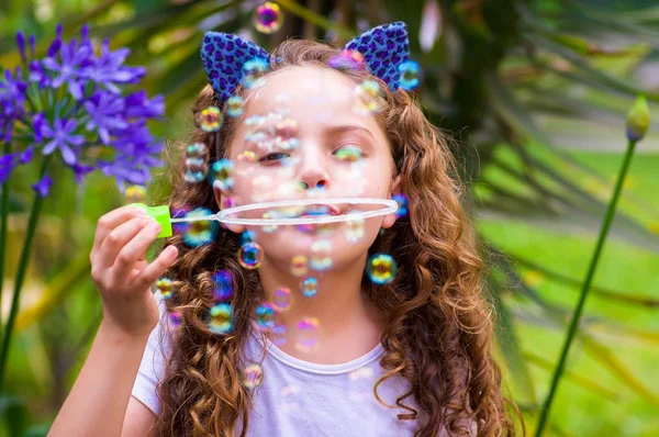 Happy little curly girl playing with soap bubbles on a summer nature, wearing a blue ears of tiger accessories over her head in a blurred nature background — Stock Photo, Image