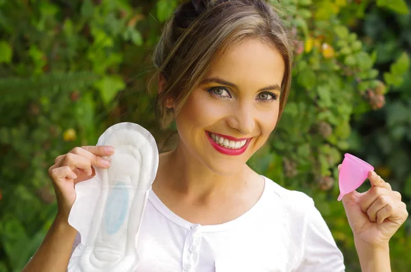 Close up van een lachende mooie jongedame die een menstruatiecup in de ene hand en een hygiënische handdoek in haar andere hand. Gynaecologie-concept, met een onscherpe achtergrond — Stockfoto