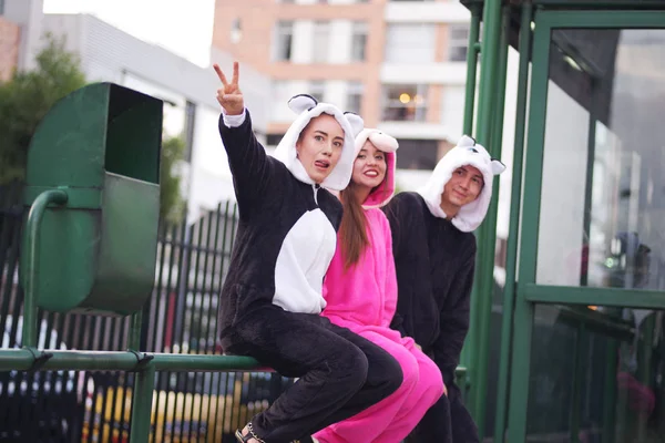 Close up de um grupo feliz de amigos, esperando o transporte público e vestindo trajes diferentes, uma mulher vestindo um traje de unicórnio rosa, outra mulher um traje de panda e o homem vestindo um gato — Fotografia de Stock