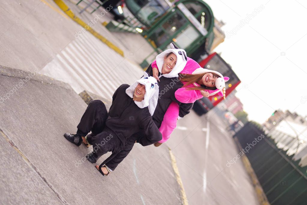 Close up of a happy group of friends, playing in the streets and wearing different costumes, one woman wearing a pink unicorn costume, other woman a panda costume and the man wearing a cat costume, in