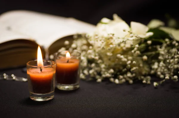 Nahaufnahme zweier orangefarbener Kerzen mit einem verschwommenen Rosenkranz über einer heiligen Bibel und kleinen Blumen, schwarzer Hintergrund — Stockfoto
