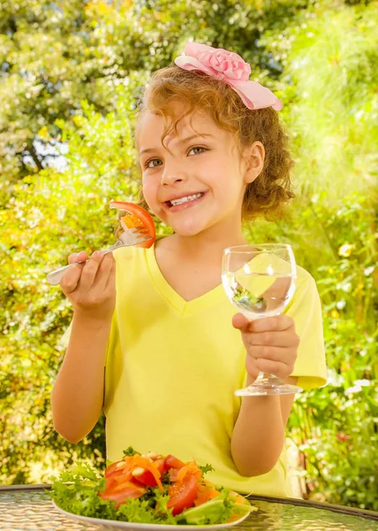 Mooi jong meisje, met een geel t-shirt een tomaat uit haar salade eten en houden van glas water, in een tuin achtergrond — Stockfoto