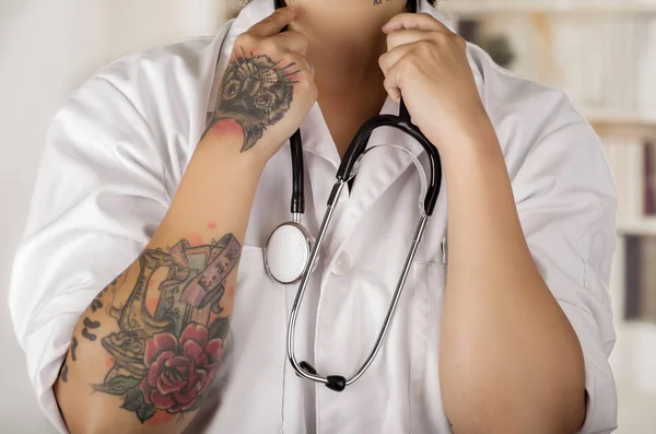 Close up of a tattooed young doctor with a stethoscope around her neck, in office background — Stock Photo, Image