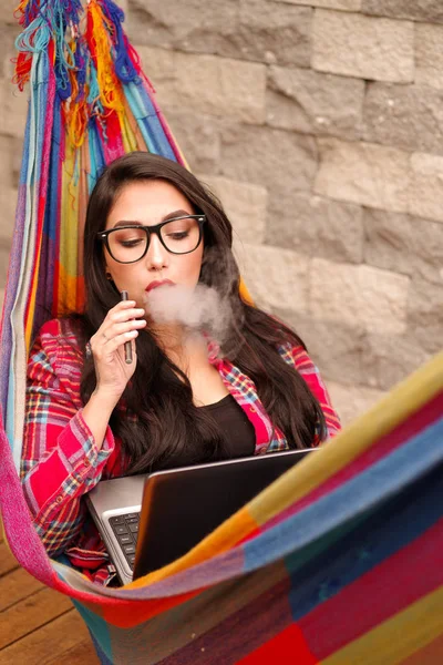 Linda hipster relaxante e deitado em uma rede colorida e desfrutar da vida, usando seu celular e computador, enquanto ela está fumando — Fotografia de Stock