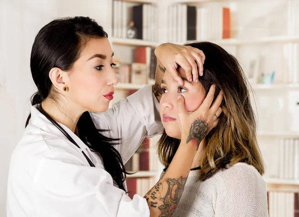 Vackra tatuerade ung läkare använder hennes hand för att kontrollera ögonen på hennes patient, i office bakgrunden — Stockfoto