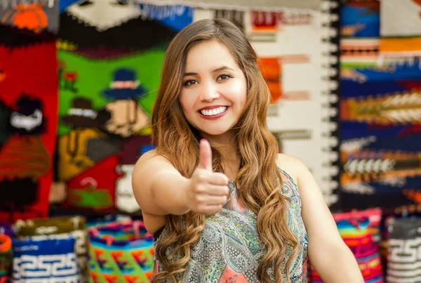 Mooie jonge vrouw met humbs lachend omhoog en poseren voor de camera, met kleurrijke Andes traditionele kleding stoffen achtergrond — Stockfoto