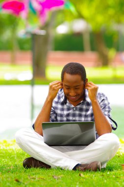 Yeşil çimenlerin üzerinde oturarak ve bilgisayarında çalışan ve onun kulaklık şehir Quito Ekvator ile müzik dinlemek adam genç siyah