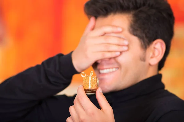 QUITO, ECUADOR - 17 OCTOBER, 2015: Close up of a smiling young man holding in his hand a golden gift present and covering his eyes with his hand, wearing a black turtleneck jacket in a blurred — стоковое фото