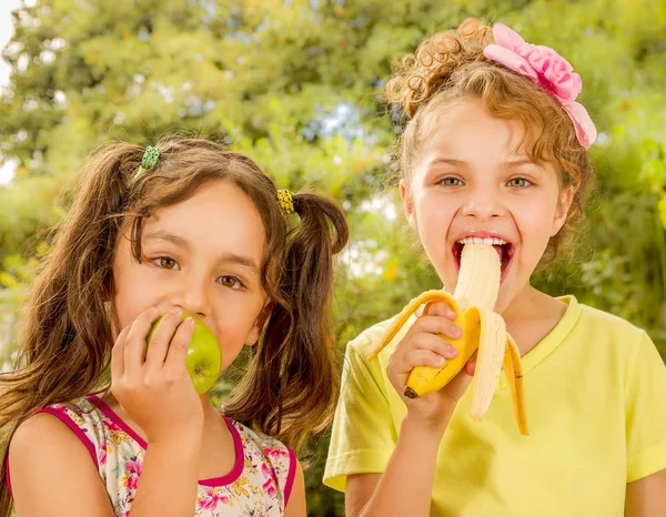 Two beautiful young girls, eating a healthy apple and banana in a garden background — Stock Photo, Image