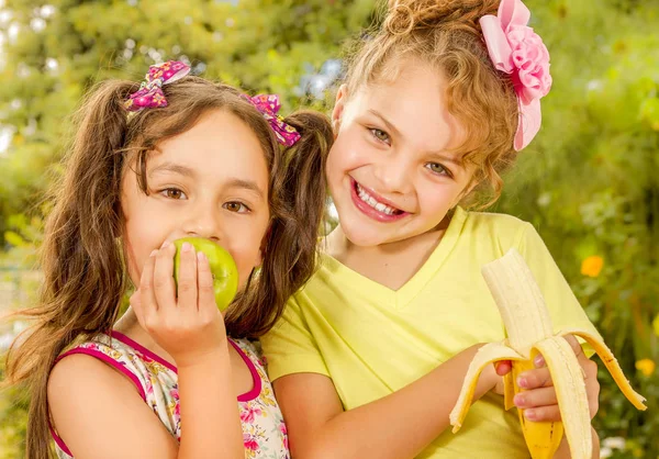 Dos hermosas chicas jóvenes, comiendo una manzana sana y plátano en un fondo de jardín —  Fotos de Stock