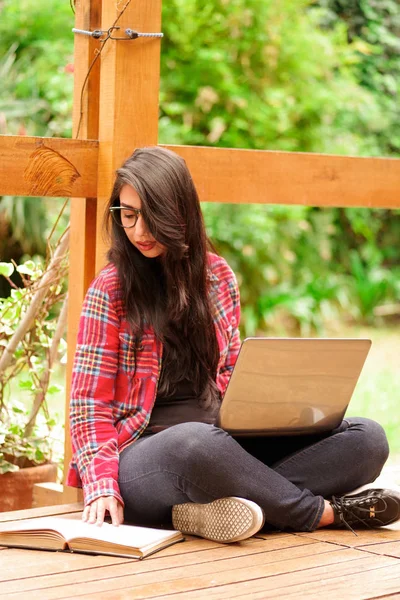 Cool lachende hipster student vrouw brillen bril. Kaukasische vrouwelijke universitaire student met een computer in haar benen, op zoek naar een boek in de achtertuin achtergrond in de buitenlucht — Stockfoto