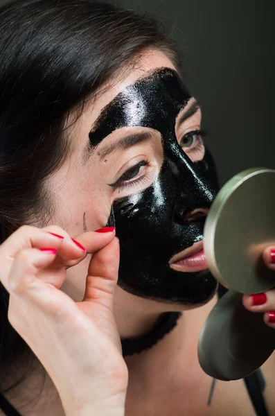 Närbild av en skönhet ung kvinna tar bort hälften av en svart ansiktsmask tittar på spegel — Stockfoto