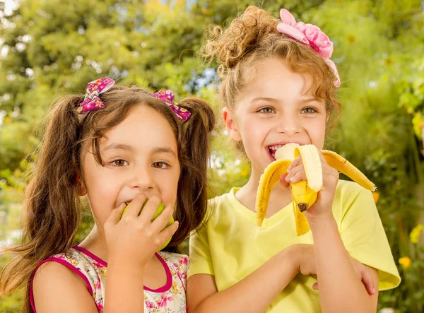 Two beautiful young girls, eating a healthy apple and banana in a garden background — Stock Photo, Image