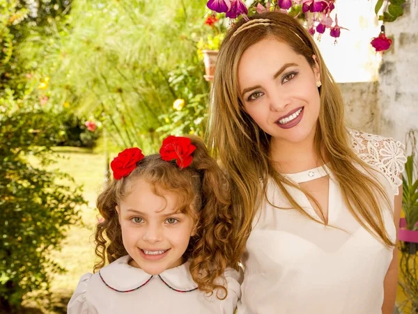 Sonriendo hermosa mamá abrazando a su bonita hija en vestido beige con dos corbatas rojas en el pelo —  Fotos de Stock