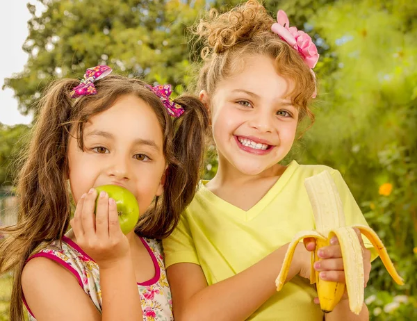 Twee mooie jonge meisjes, eten van een gezonde appel en banaan in een tuin achtergrond — Stockfoto