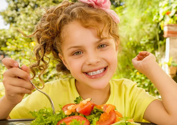 Primer plano de una hermosa niña sonriente, con una camiseta amarilla que se prepara para comer una ensalada saludable con un tenedor, en un fondo de jardín —  Fotos de Stock