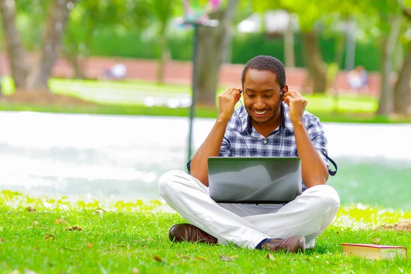 Yeşil çimenlerin üzerinde oturarak ve bilgisayarında çalışan ve onun kulaklık şehir Quito Ekvator ile müzik dinlemek adam genç siyah