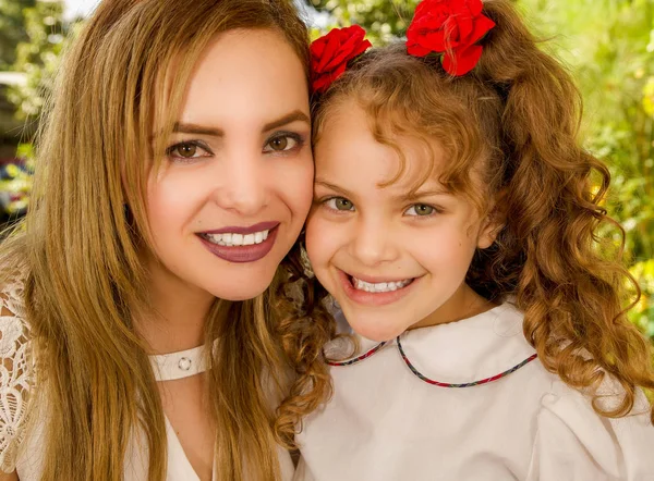 Close up of a smiling beautiful mom hugging her pretty daugher in beige dress wearing two red ties in hair — Stock Photo, Image