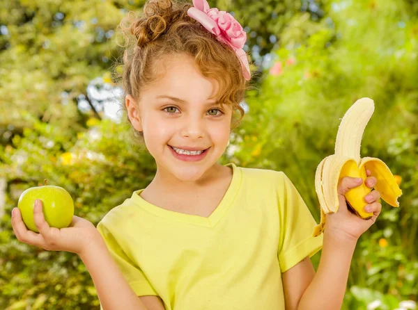 Όμορφη νεαρή κοπέλα, φοράει ένα κίτρινο μπλουζάκι κρατώντας ένα υγιές μήλο και μπανάνα, σε φόντο στον κήπο — Φωτογραφία Αρχείου