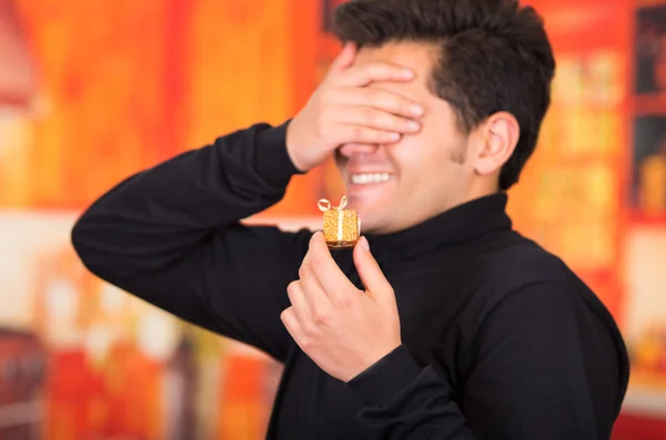 QUITO, EQUADOR - 17 DE OUTUBRO DE 2015: Fechar um jovem sorridente segurando na mão um presente de ouro e cobrindo os olhos com a mão, usando uma jaqueta preta de gola alta em uma embaçada — Fotografia de Stock