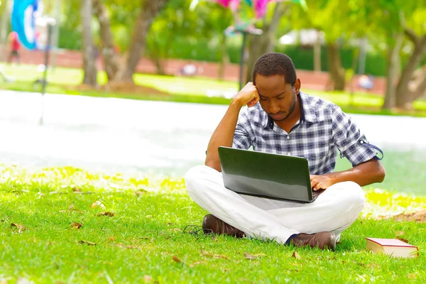 Joven hombre negro sentado sobre hierba verde y trabajando en su computadora en la ciudad de Quito Ecuador — Foto de Stock