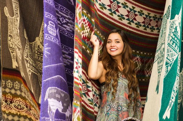 Belle jeune femme souriante touchant les vêtements traditionnels andins et posant pour appareil photo, fond de tissus colorés — Photo
