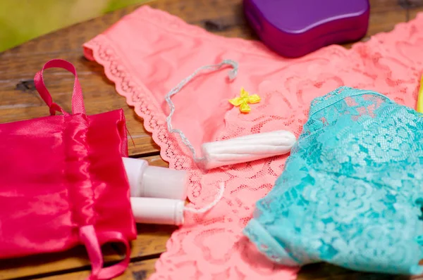 Primer plano de un tampón de algodón de menstruación sobre una ropa interior de mujer rosa y azul, y un bolso de algodón rojo, en una mesa de madera, y una caja de plástico púrpura, en un fondo borroso —  Fotos de Stock