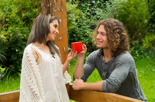 Lyckliga par i kärlek vid utomhus och ler, man delar hans kopp kaffe till hennes flickvän en tittar hennes flickvän, i en uteplats bakgård bakgrund — Stockfoto