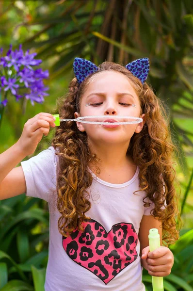Blij krullend meisje spelen met zeepbellen op een zomer natuur, het dragen van een blauwe oren van tijger accessoires over haar hoofd op een achtergrond wazig natuur — Stockfoto