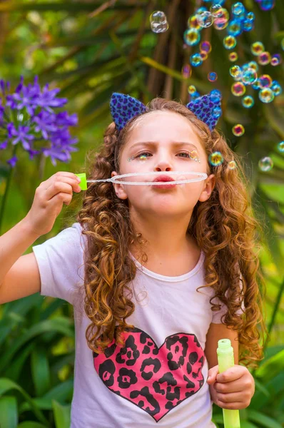 Happy little curly girl playing with soap bubbles on a summer nature, wearing a blue ears of tiger accessories over her head in a blurred nature background