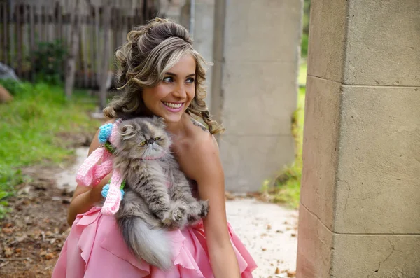 美丽微笑的女人，身穿粉红色连衣裙的公主拥抱她的猫，在室外 — 图库照片