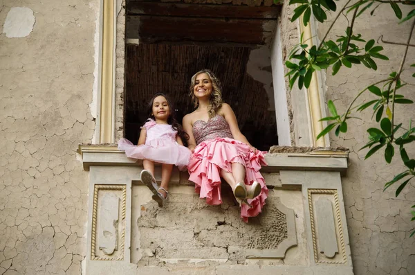 Schöne Frau und ihre süße kleine Tochter lächeln und blicken in die Kamera, sitzen im Fenster eines Schlosses und tragen ein rosafarbenes Prinzessinnenkleid — Stockfoto