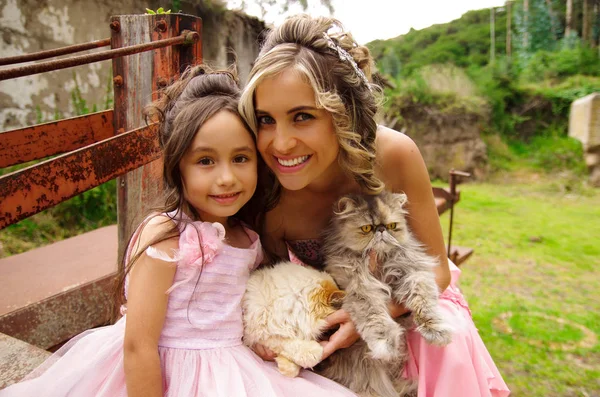 Nahaufnahme einer schönen Frau und ihrer niedlichen kleinen Tochter, die in die Kamera schaut und lächelt, in einem Prinzessinnenkleid, während sie ihre Katzen im Freien umarmen — Stockfoto