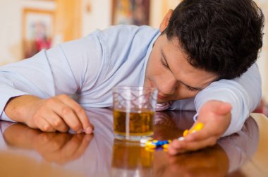 Ahşap bir masa üzerinde döşeme ve onlarca hapları elinde ve onun diğer el, aşırı doz veya intihar alkollü içki tutan genç adam