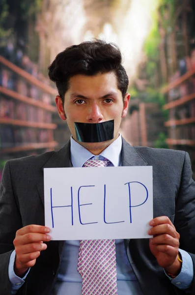 Empresario con una cinta negra en la boca y sosteniendo en la mano un cartel blanco con una ayuda, en un fondo borroso — Foto de Stock