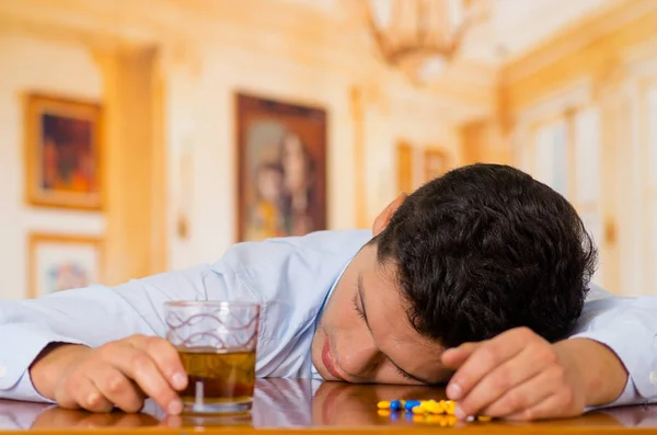 Junger Mann, der sich über einen Holztisch legt und Dutzende Tabletten in der Hand hält und in der anderen Hand ein alkoholisches Getränk, Überdosis oder Selbstmord — Stockfoto