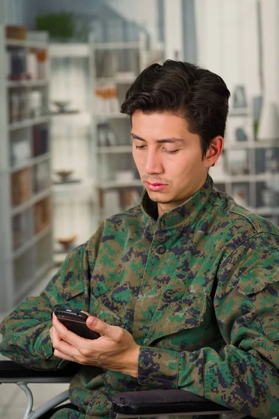 Nahaufnahme eines hübschen jungen Soldaten, der im Rollstuhl sitzt und sein Handy benutzt, vor verschwommenem Hintergrund — Stockfoto